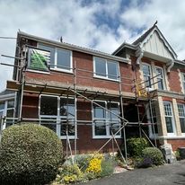 A brick house with white trim and large windows is partially covered by domestic scaffolding, indicating construction or renovation work. A garden with bushes and flowers sits in front, beneath a partly cloudy sky.