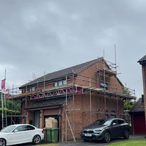 A brick house with commercial scaffolding around the upper level, indicating construction or renovation work. Two cars are parked in the driveway under a cloudy, gray sky.