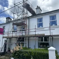 A white two-story house with domestic scaffolding set up in front, reaching above the roof. The sky is partly cloudy, and there are green plants and a white fence in the foreground.