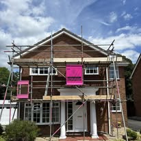 A two-story brick house with white columns is surrounded by domestic scaffolding marked by pink signs, under a partly cloudy sky. Renovation or construction work appears to be in progress.