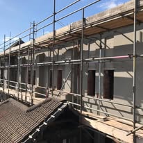 A building under construction or renovation with Domestic Scaffolding and scaffolding set up along its exterior wall. Several windows are visible, and the sky is clear with some clouds.
