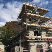 A building under construction, covered in commercial scaffolding, with workers visible on the upper levels. The sky is partly cloudy and trees are visible in the background.