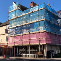 A three-story building covered in scaffolding and blue and pink protective netting, with temporary roofs overhead, is undergoing construction or renovation under a clear blue sky.