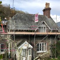 A stone cottage with a tiled roof is surrounded by scaffolding for construction or repairs. A sign on the scaffolding highlights experienced scaffolders, while chimney wraps are visible against trees and a cloudy sky.