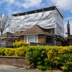A brick house with a tiled roof is covered in commercial scaffolding and white sheeting, likely for renovation or roof work. The front yard is lined with green bushes, and temporary roofs provide added protection while work is underway.