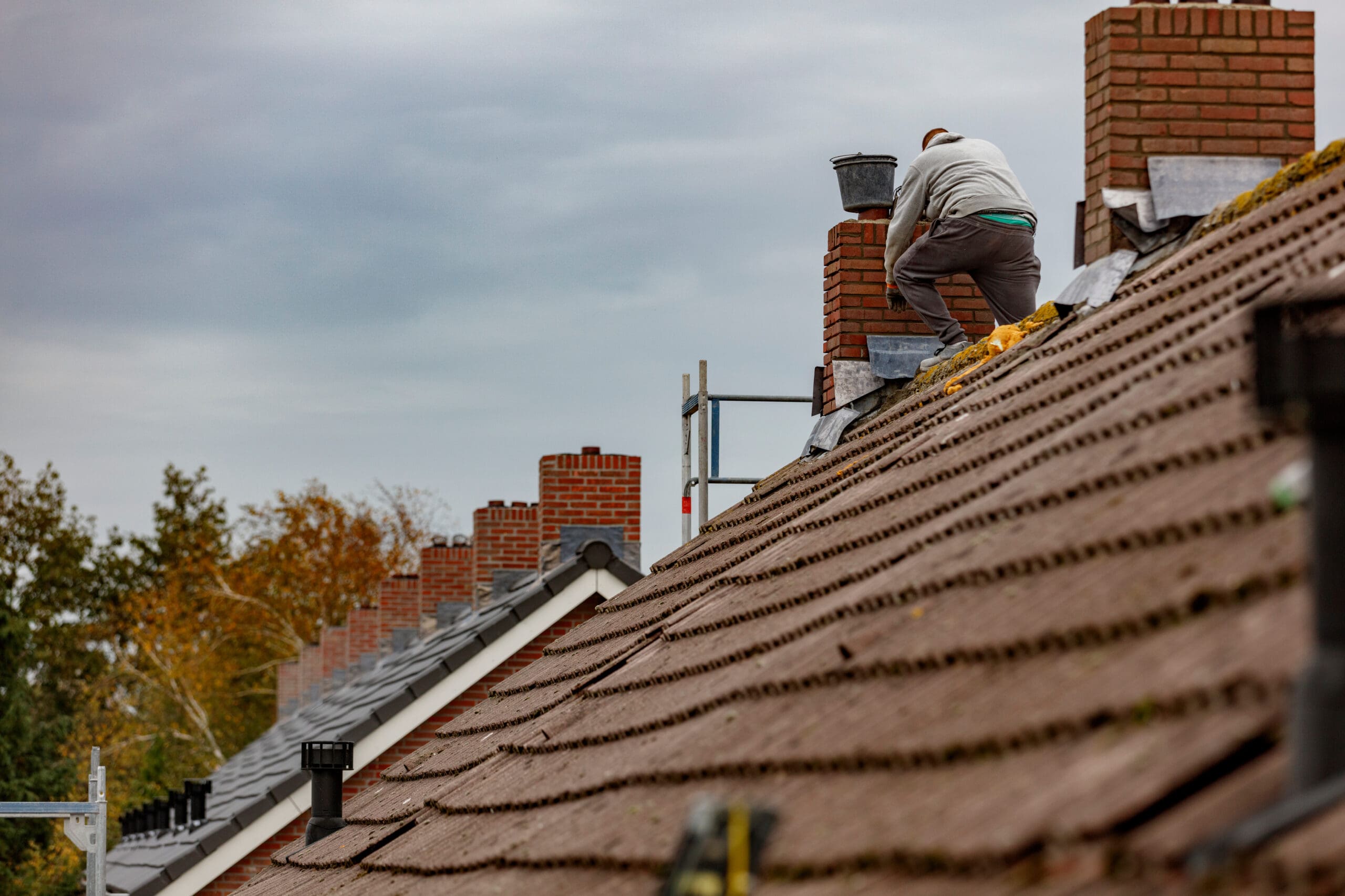 A person in work clothes crouches on a sloped, tiled roof, repairing a brick chimney with chimney wraps. Other chimneys and rooftops are visible in the background beneath a cloudy sky.