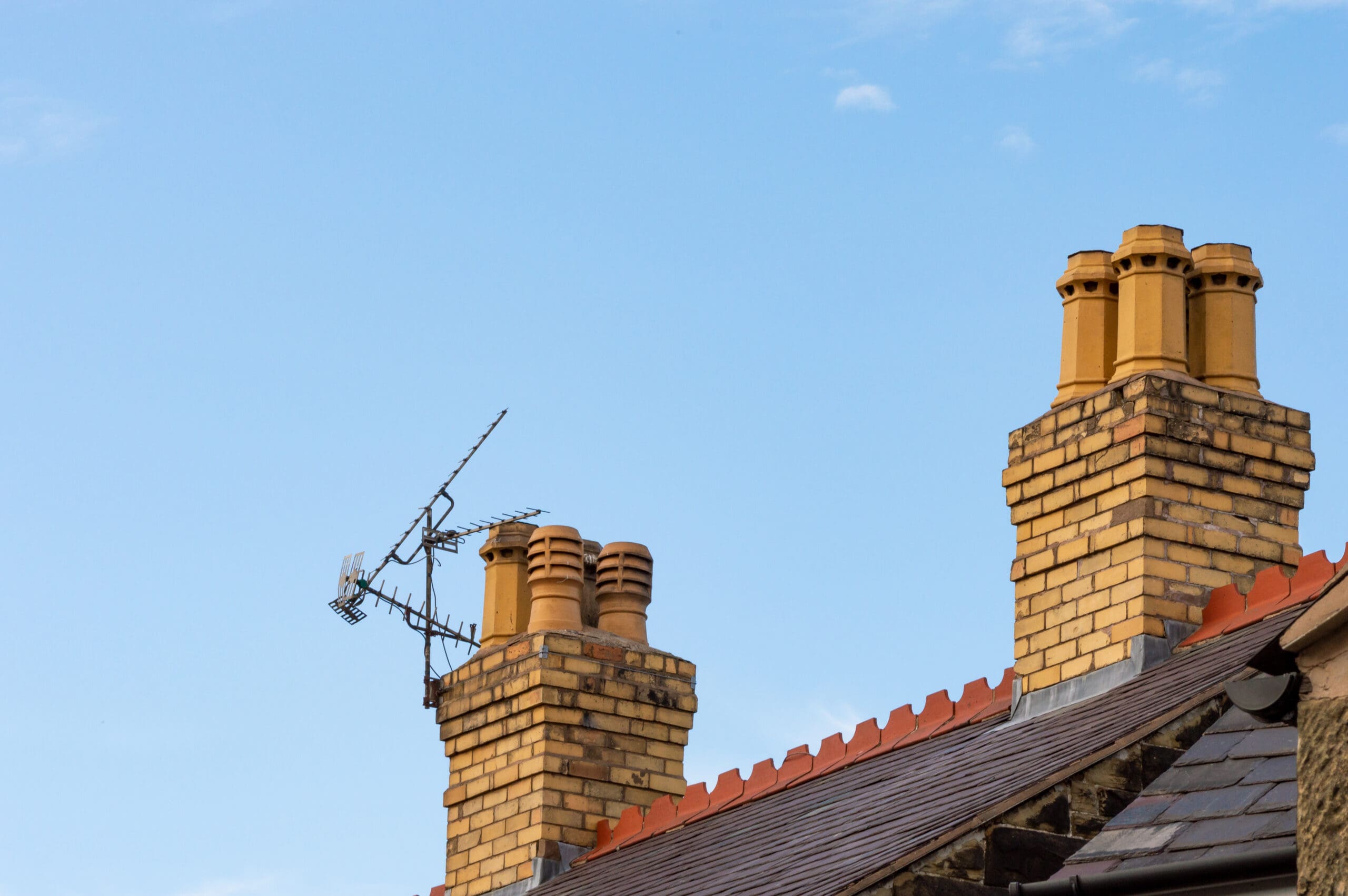 Two brick chimneys with multiple clay chimney pots on a slate rooftop, one fitted with a TV antenna. The clear blue sky forms the background—ideal conditions for a scaffolder to install domestic scaffolding or chimney wraps.