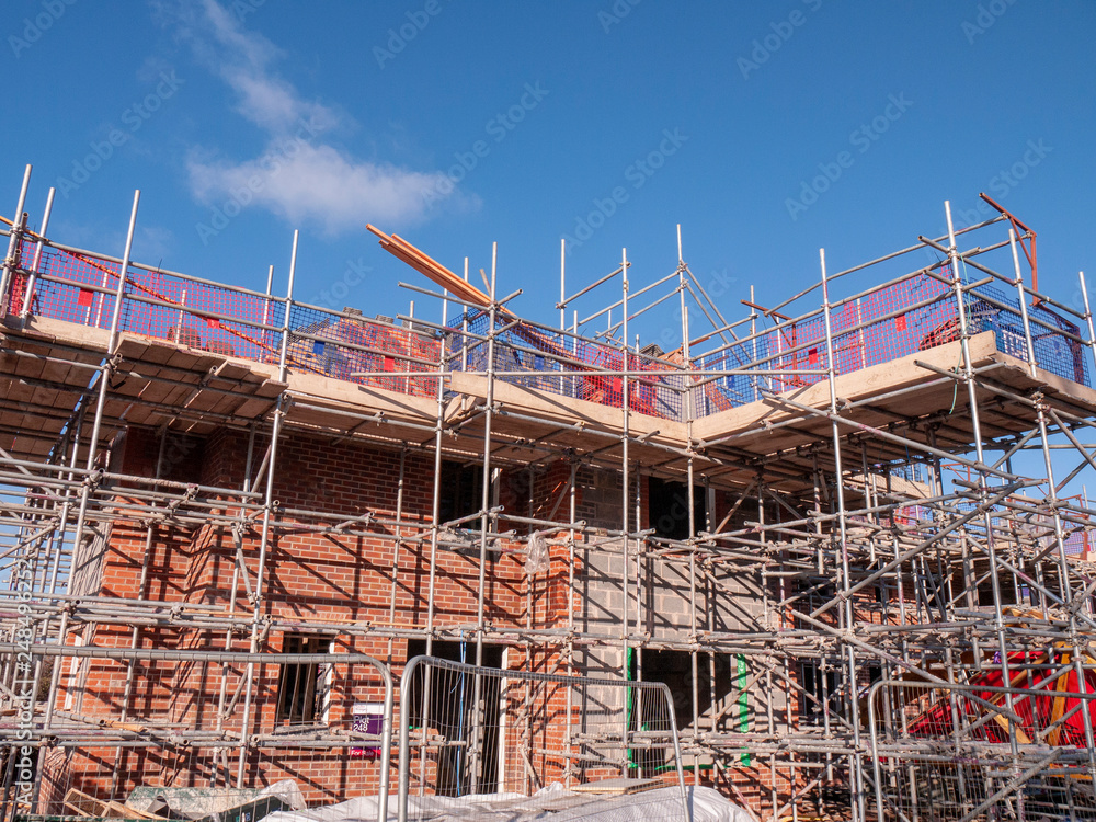 Domestic scaffolding surrounds a partially constructed brick building under a clear blue sky, with safety nets and construction materials visible on the upper level.