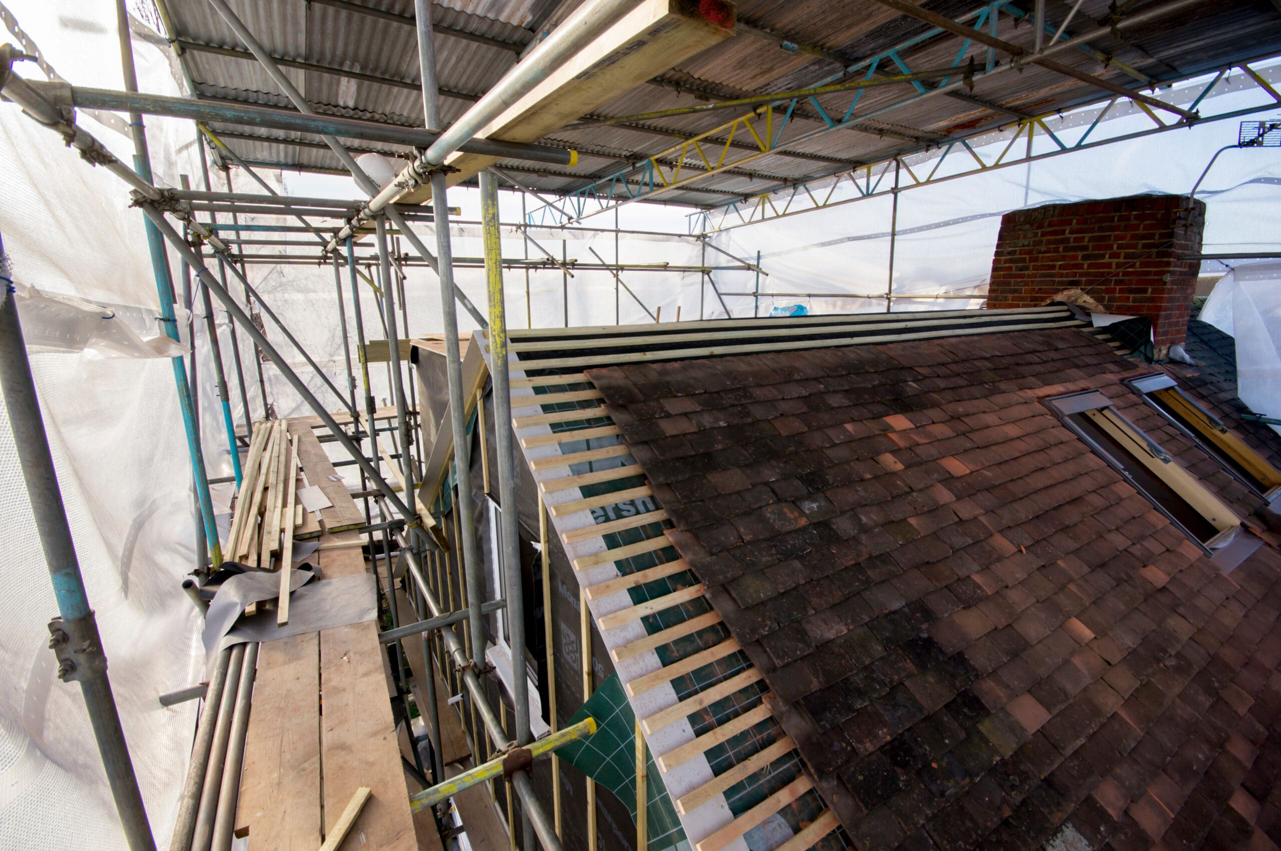 A roof under renovation is partially covered with wooden battens and roof tiles. Commercial scaffolding and protective sheeting surround the structure, with construction materials and tools visible on the scaffolding platform.