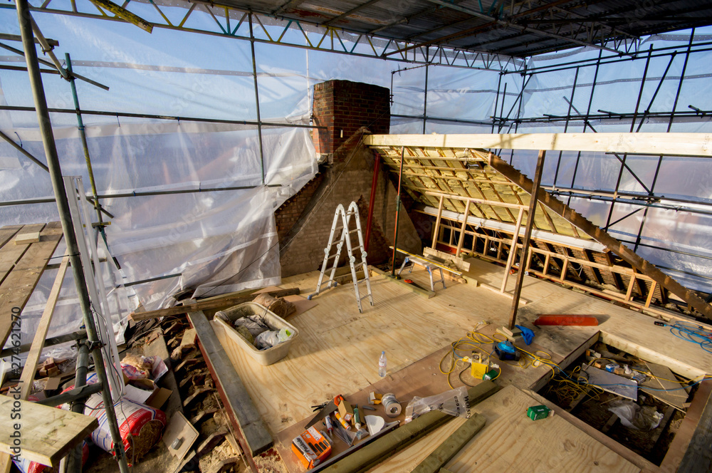 A construction site on a rooftop features Commercial Scaffolding, a partially built wooden structure, tools, building materials, and a metal ladder near a brick chimney under a transparent plastic covering.