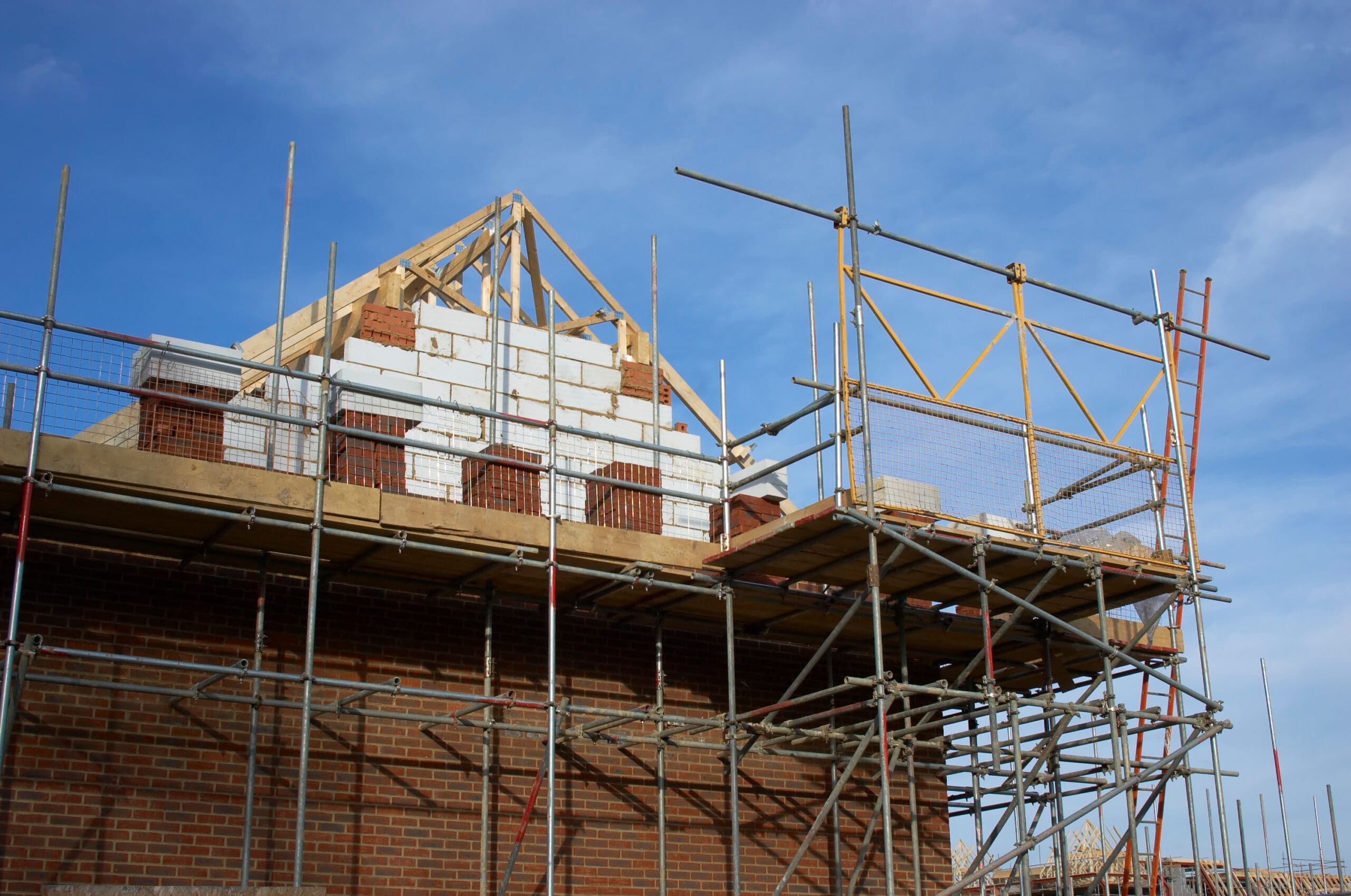 A partially built brick house with wooden roof framing, white blocks, and commercial scaffolding surrounds the structure under a blue sky.