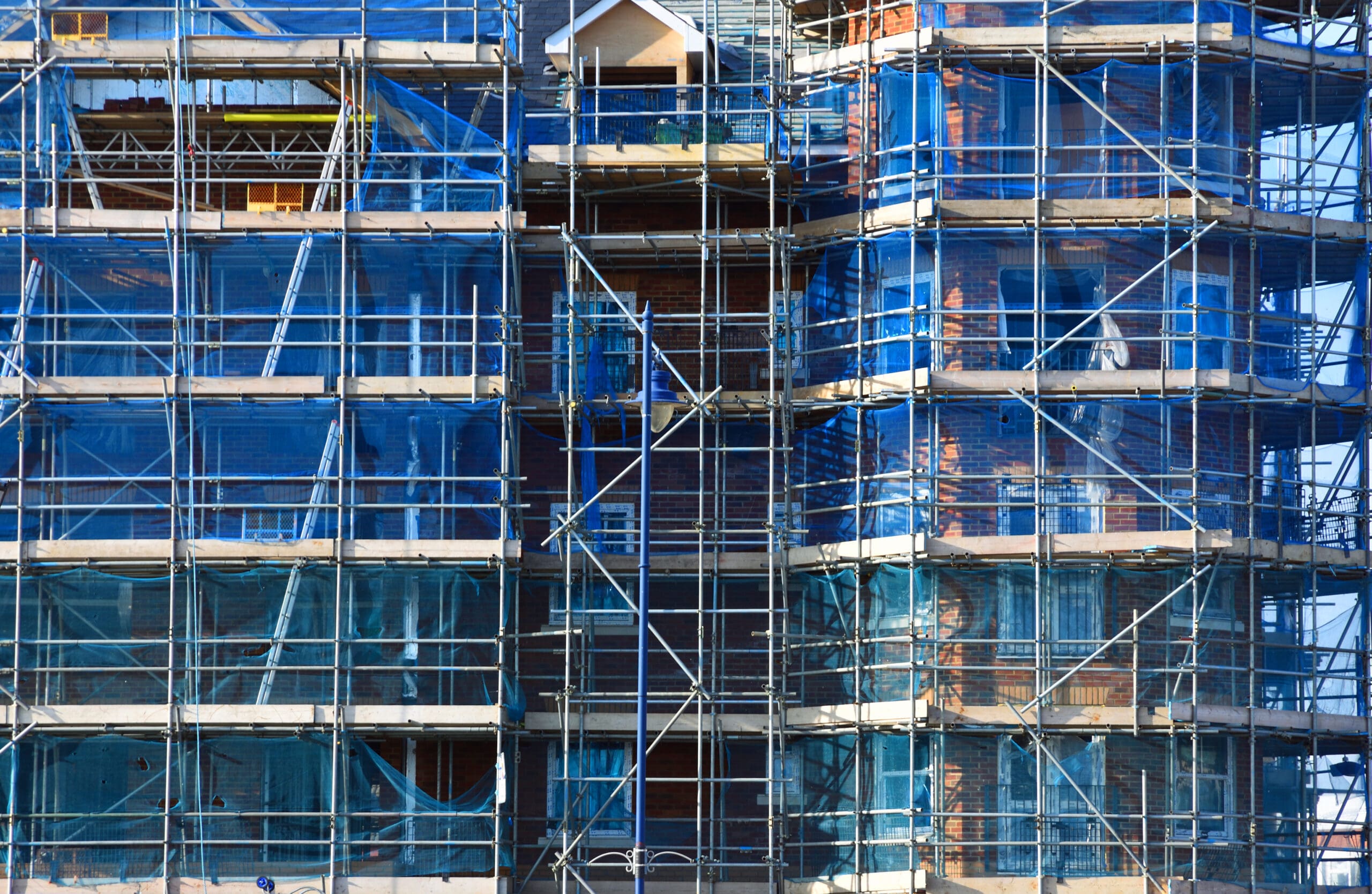 A multi-story building under construction, covered with blue safety netting and surrounded by domestic scaffolding. The structure has exposed brick walls and partially finished sections.