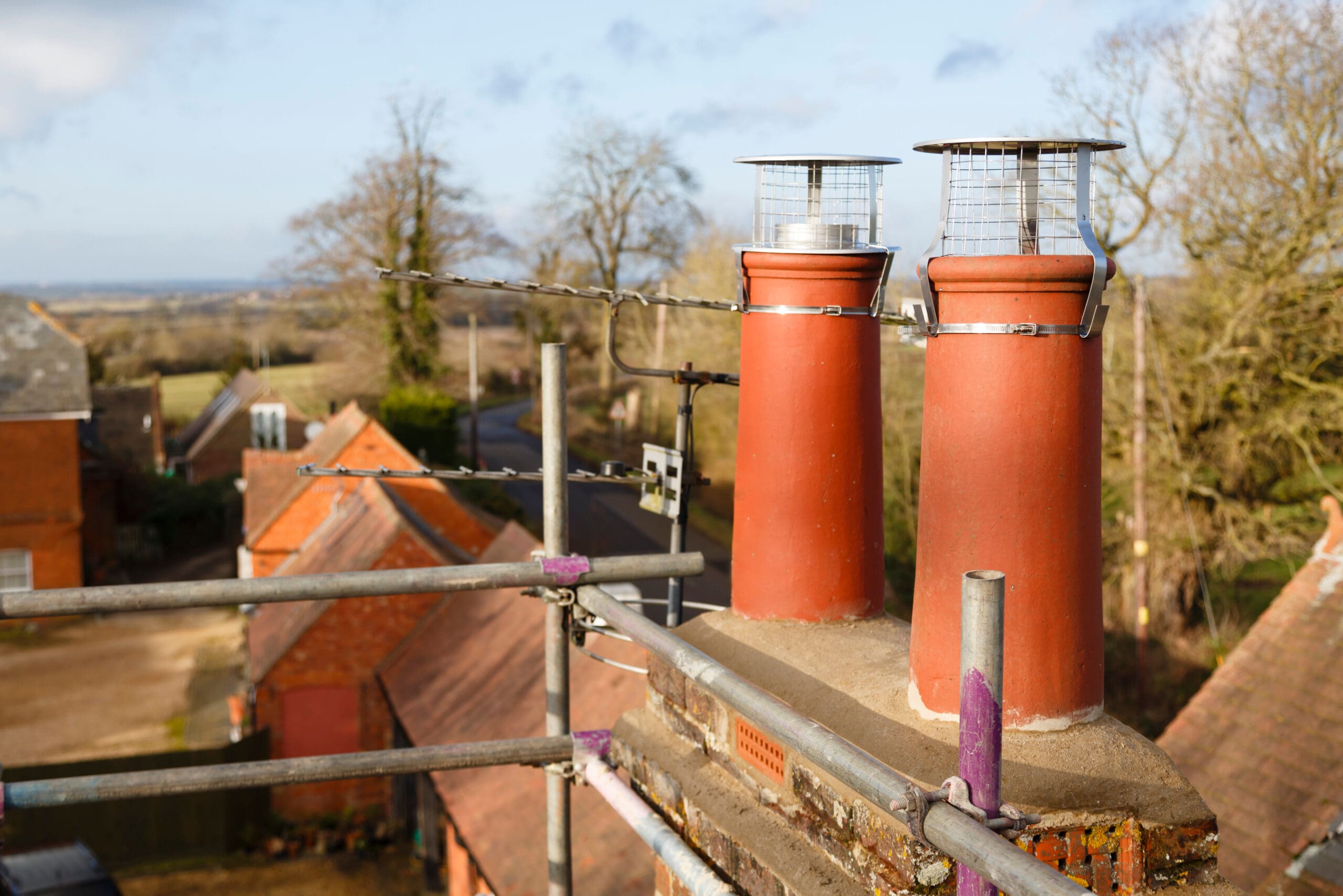 Two red chimney pots with metal caps on a rooftop, seen from above. Commercial scaffolding surrounds the chimneys, and a village with houses, trees, and a winding road is visible in the background under a blue sky.
