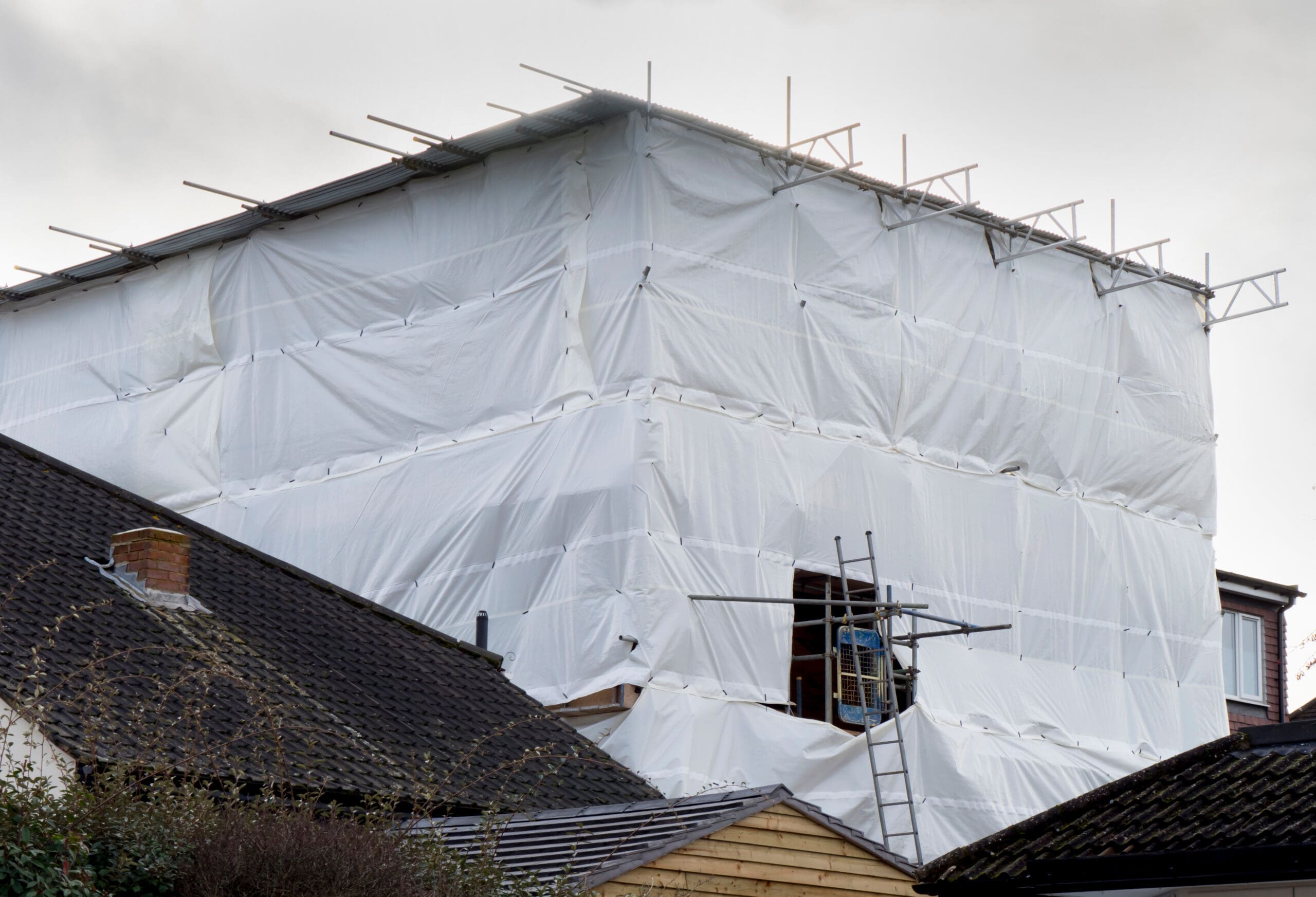 A large building under construction or renovation, covered in white plastic sheeting and commercial scaffolding, rises above the rooftops of neighboring houses on a cloudy day.