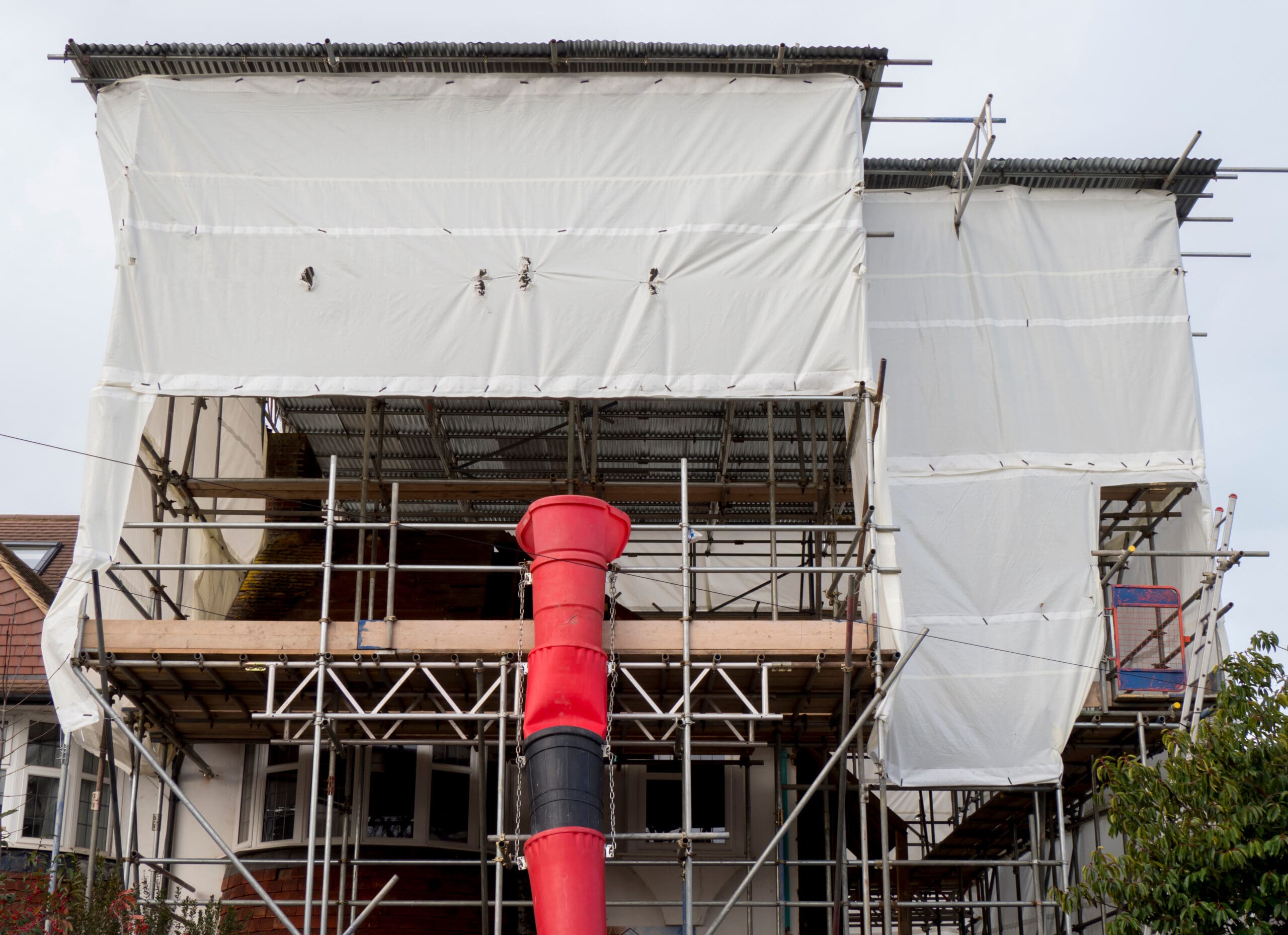 A house under construction is covered in domestic scaffolding and white protective sheeting, with a red construction chute attached to the upper level. Some windows and parts of the structure, including chimney wraps, are visible behind the sheeting.