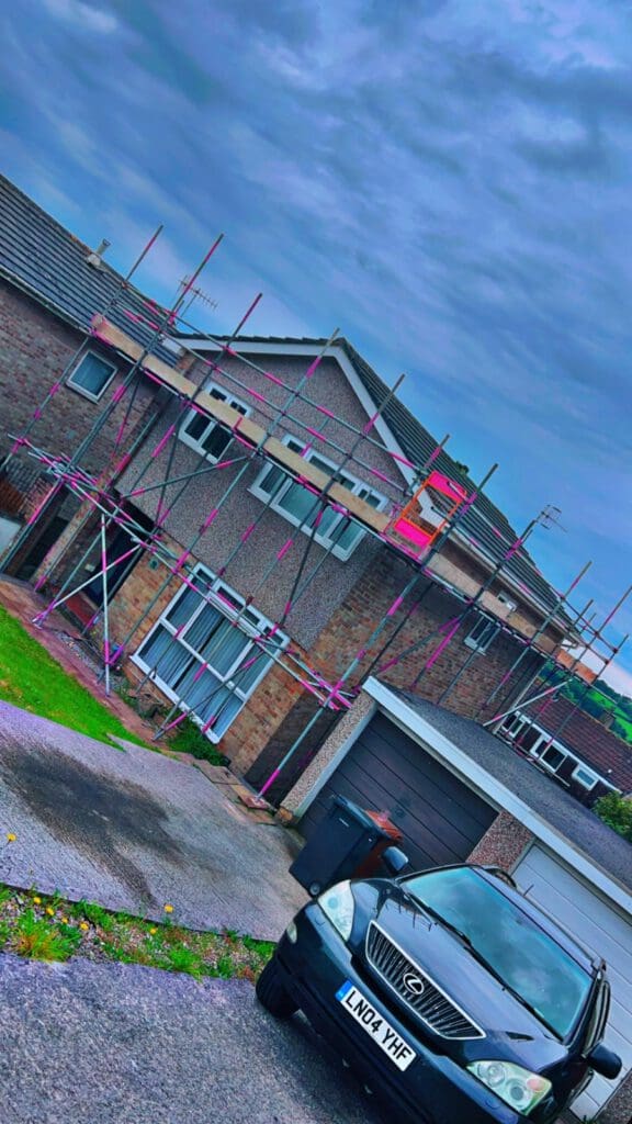 A two-story house with brick walls and a garage is under construction, surrounded by pink domestic scaffolding. A black Lexus car with the license plate LN04 YHF is parked in the driveway under a cloudy sky.