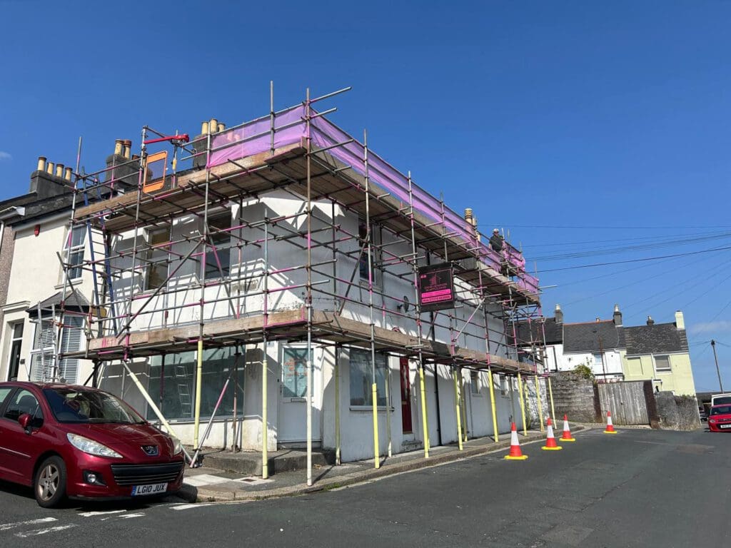 A two-story white building covered in commercial scaffolding stands on a street corner under a clear blue sky. Orange traffic cones and parked cars line the street as scaffolders work beneath temporary roofs.