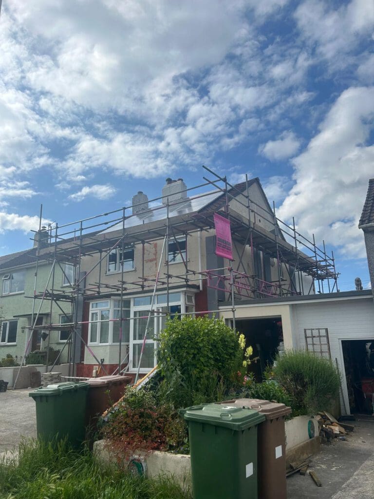 A two-story house with domestic scaffolding and temporary roofs around the exterior, suggesting renovation or construction work. The sky is partly cloudy, and green bins are lined up in front of the driveway and a small garden.