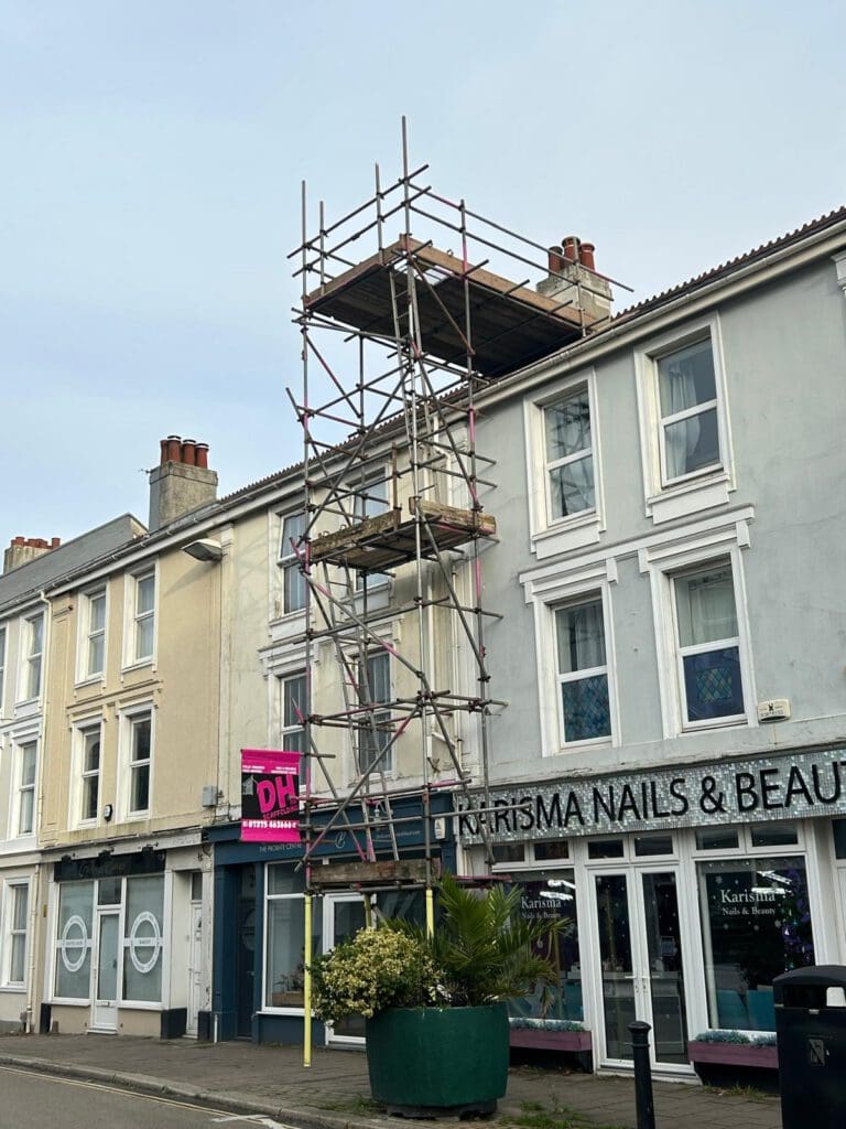 Tall commercial scaffolding stands on the sidewalk in front of a row of shops, including a nail and beauty salon. The scaffolder’s work reaches above the rooftops, while a palm plant in a large green pot sits nearby.