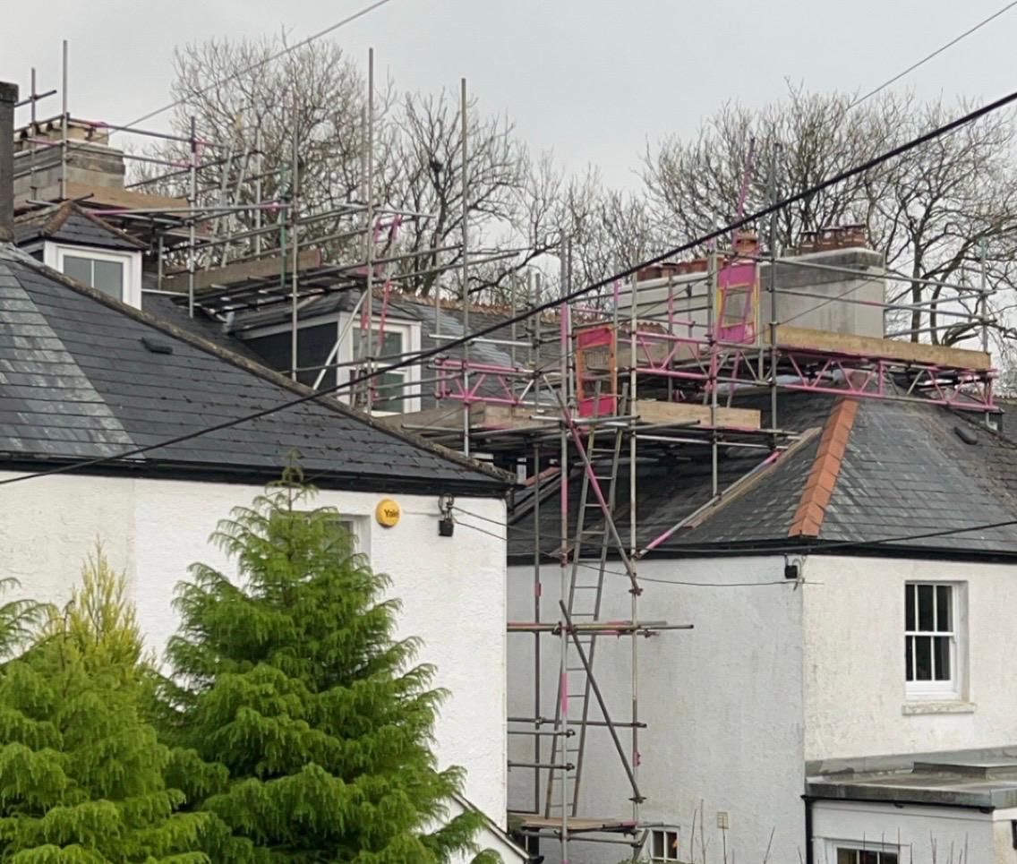 Domestic scaffolding covers the roofs of white houses under renovation, with construction equipment visible. Leafless trees stand in the background while green shrubs line the foreground. Overhead wires cross the scene.