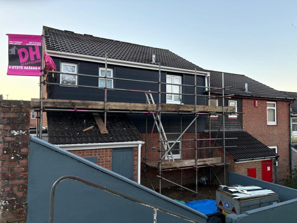 A two-story brick house under renovation with domestic scaffolding and temporary roofs along the upper floor. A pink DH Carpentry banner hangs on the scaffolding, while construction materials and tools are visible in the yard below.
