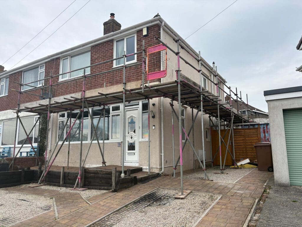 A two-story semi-detached house with domestic scaffolding and a temporary roof around the front and side, indicating construction or renovation work. The driveway is paved, and a green garage door is visible to the right.