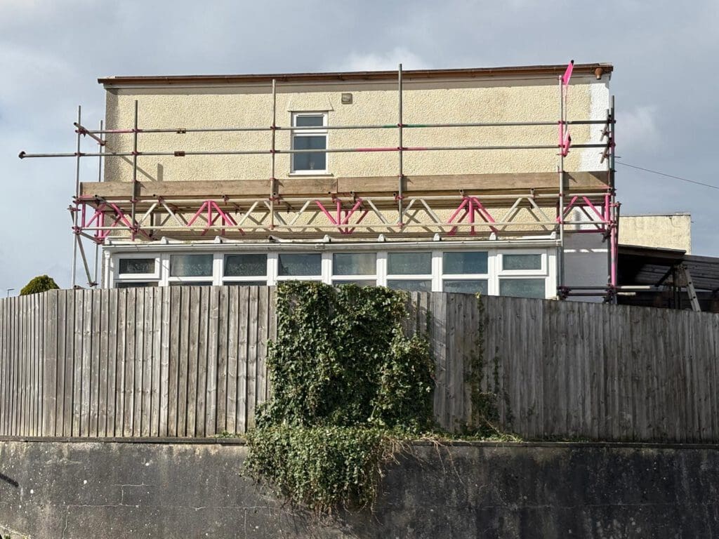 A two-story house with scaffolding and a temporary roof on the upper floor, partially obscured by a tall wooden fence covered in ivy. The sky is overcast.