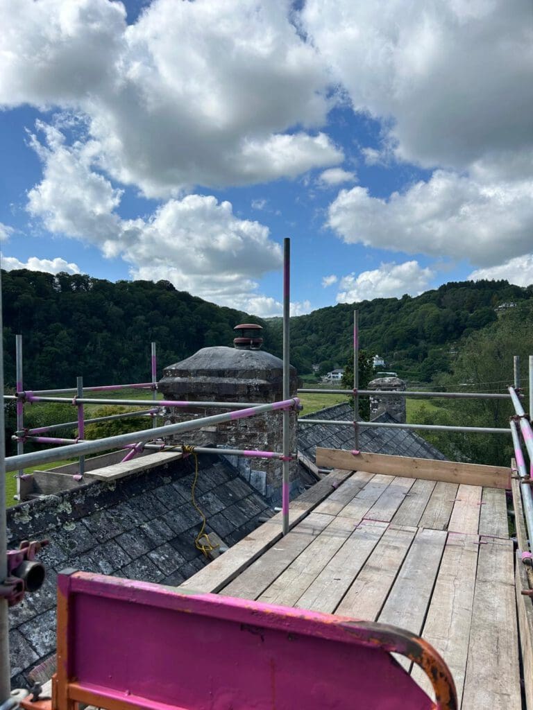A wooden scaffold with pink rails, part of a temporary roof setup, is set up on a rooftop overlooking a green, hilly landscape under a partly cloudy sky. The scaffold provides access near a chimney, with rooftops visible below.