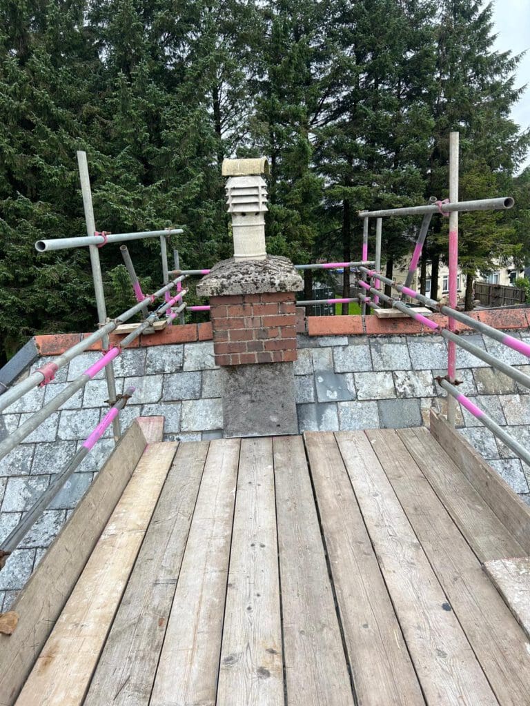 A brick chimney with a stone cap stands on a shingled roof, surrounded by domestic scaffolding and wooden planks. Tall evergreen trees are visible in the background.