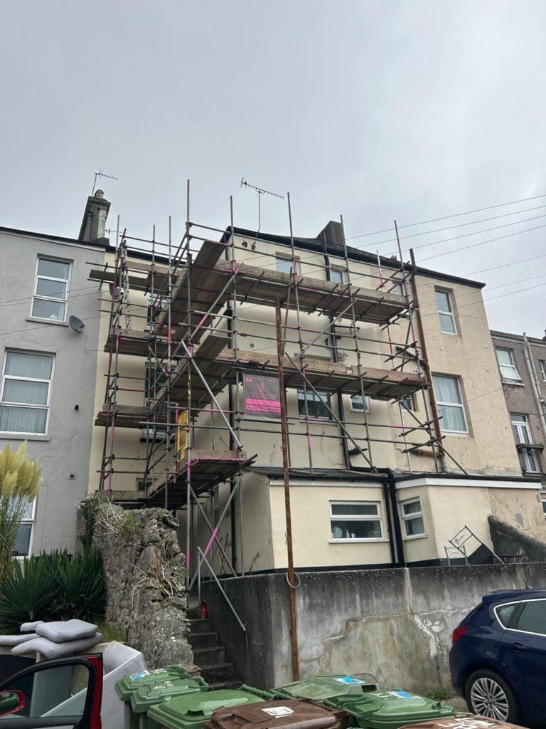 A three-story building under renovation is covered with domestic scaffolding. Green bins and a parked black car sit in front, beneath a cloudy sky. The scaffolder wrapped pink tape around parts of the scaffolding for visibility.
