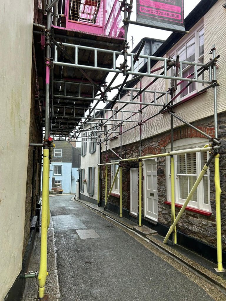 A narrow street lined with houses, where domestic scaffolding with yellow and pink parts covers one side of the road and extends overhead, leaving a small path for pedestrians. The wet road suggests recent rain.