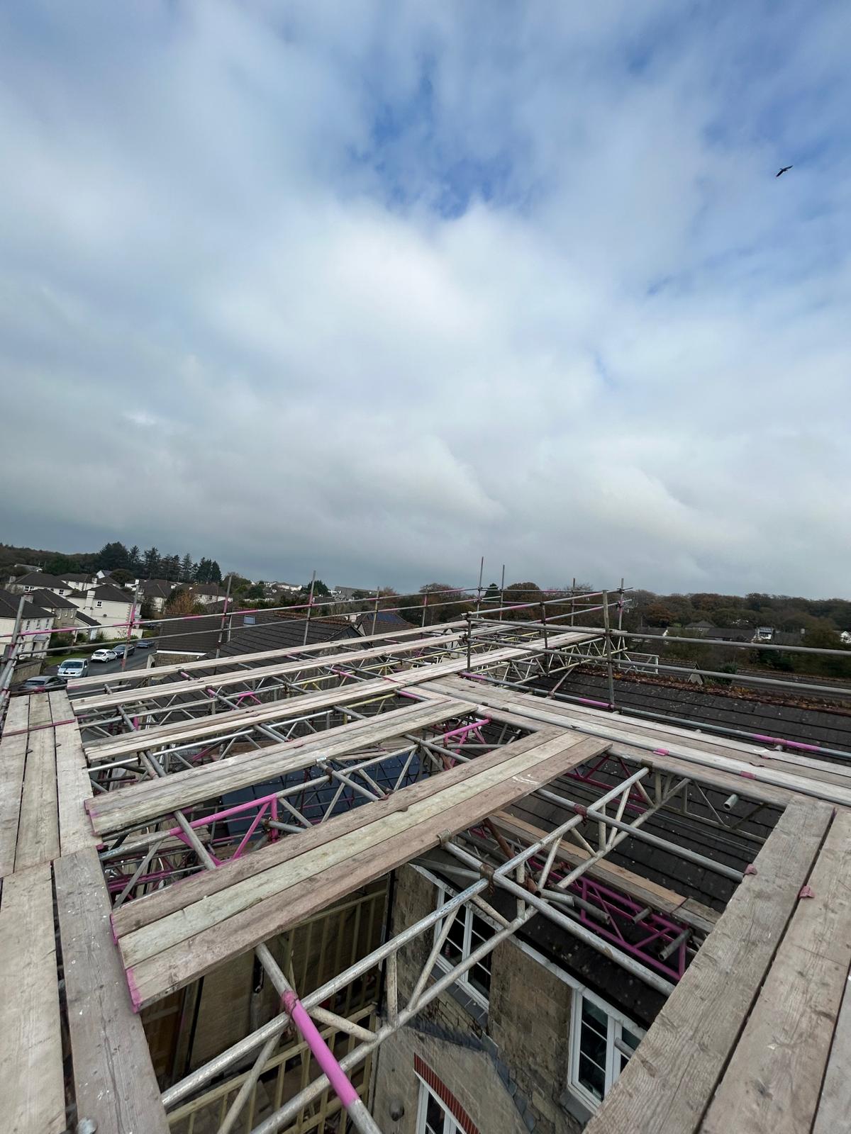 Wooden and metal commercial scaffolding, including temporary roofs, is set up around the roof of a building under construction or renovation, with views of neighboring houses, trees, and a cloudy sky in the background.
