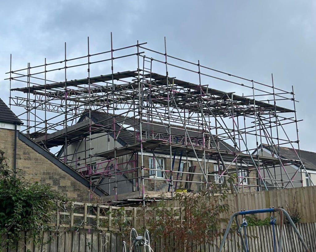 A partially constructed house surrounded by extensive domestic scaffolding, with a wooden fence and garden plants in the foreground under a cloudy sky.