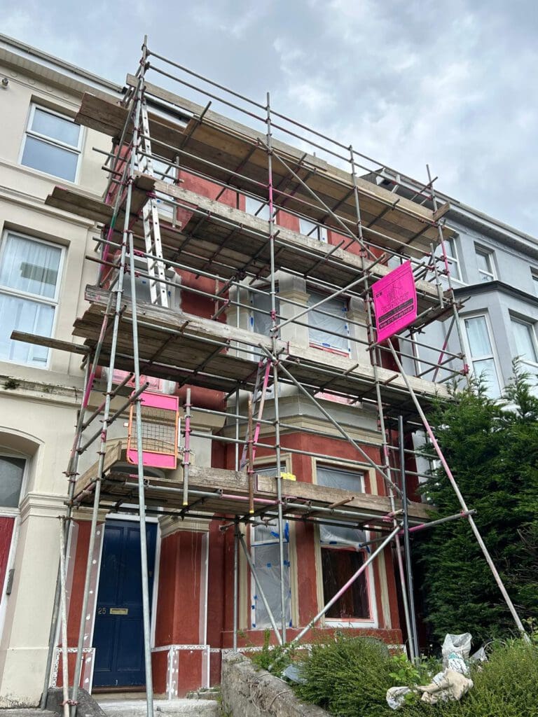 A three-story residential building with red walls is covered in domestic scaffolding and bright pink warning signs. The scaffolding reaches the temporary roof, while a tree and pathway are visible out front under a cloudy sky.