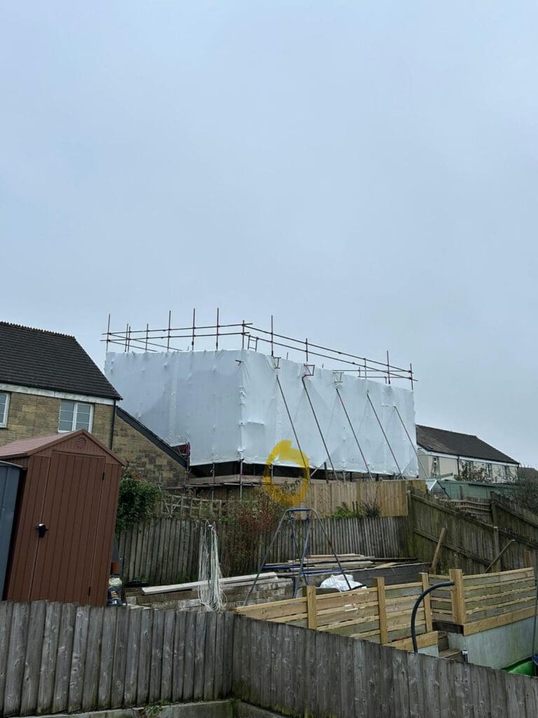 A house is covered in white scaffolding and plastic sheeting, showcasing expert domestic scaffolding with a yellow circular object on the front. The scene includes a fenced yard, a brown shed, and a cloudy sky.