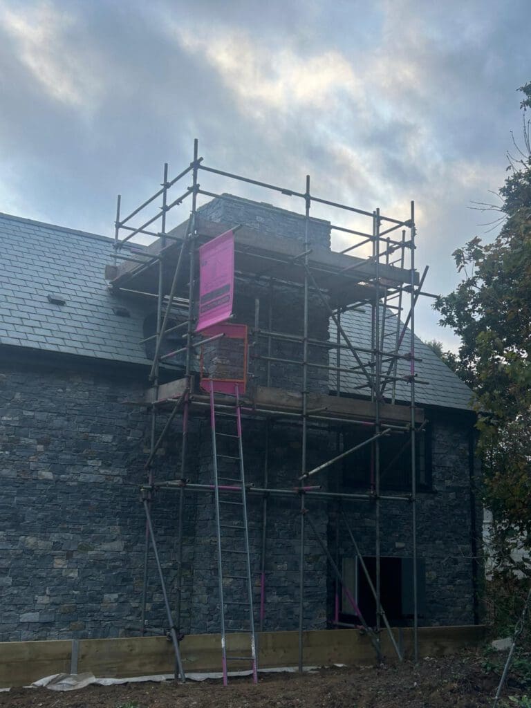A stone building with a pitched slate roof is surrounded by domestic scaffolding focused on the chimney. The sky is cloudy, and a tree is visible on the right side of the image.