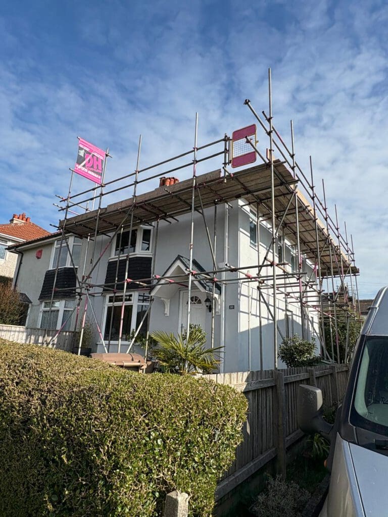 A two-story house with domestic scaffolding and temporary roofs erected around the roof for construction or repairs, under a partly cloudy sky. Two construction banners hang from the scaffolding, and a hedge lines the front yard.