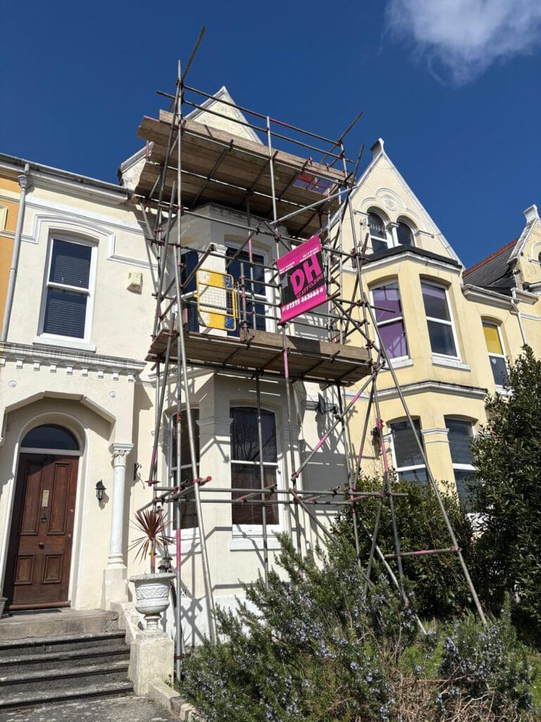 A two-story cream and yellow house with domestic scaffolding and a temporary roof set up at the front. A pink sign on the scaffolding reads "DH RESTORATIONS." The sky is blue with a few clouds overhead.