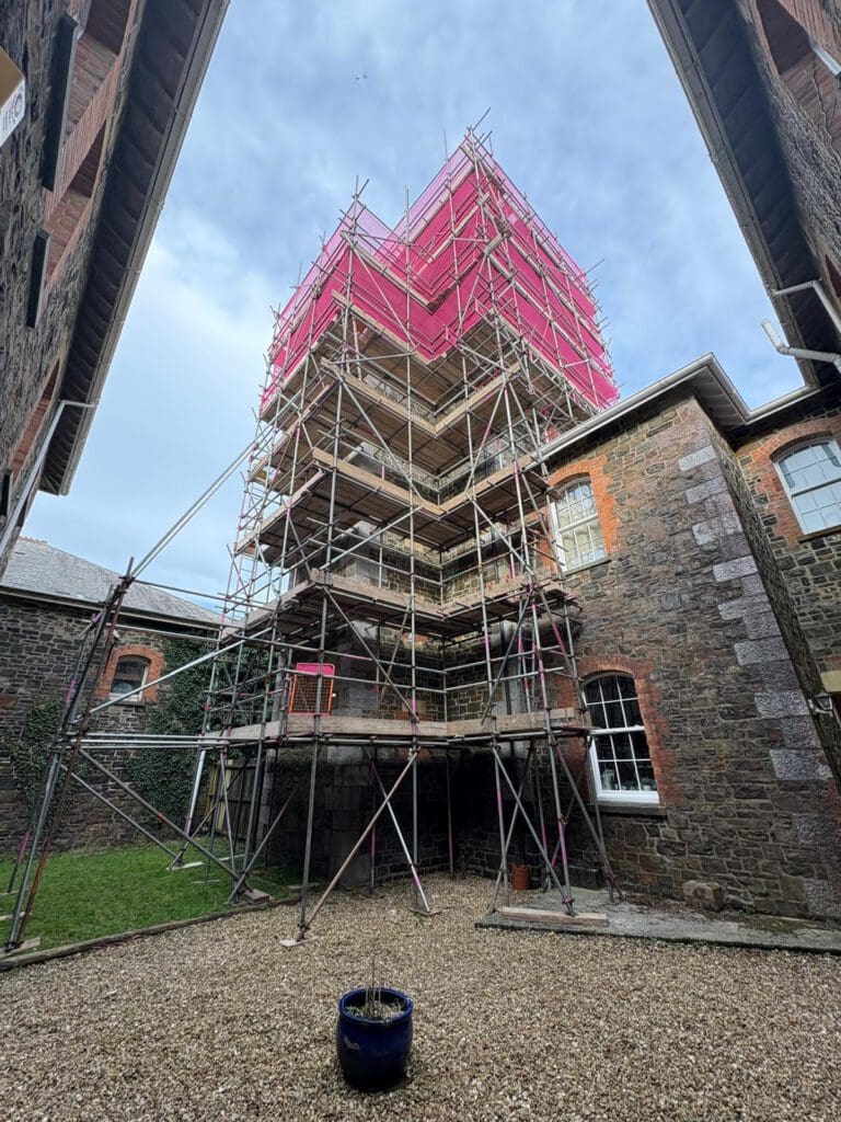 A tall scaffold topped with temporary roofs and covered with pink netting stands in a stone courtyard between two old brick buildings under a blue sky. A blue planter with a bare plant sits on gravel in the foreground.