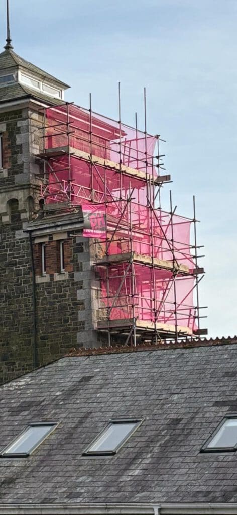 A stone building tower is surrounded by pink construction scaffolding and temporary roofs. The sky is clear and blue, with part of a roof featuring skylights visible in the foreground.