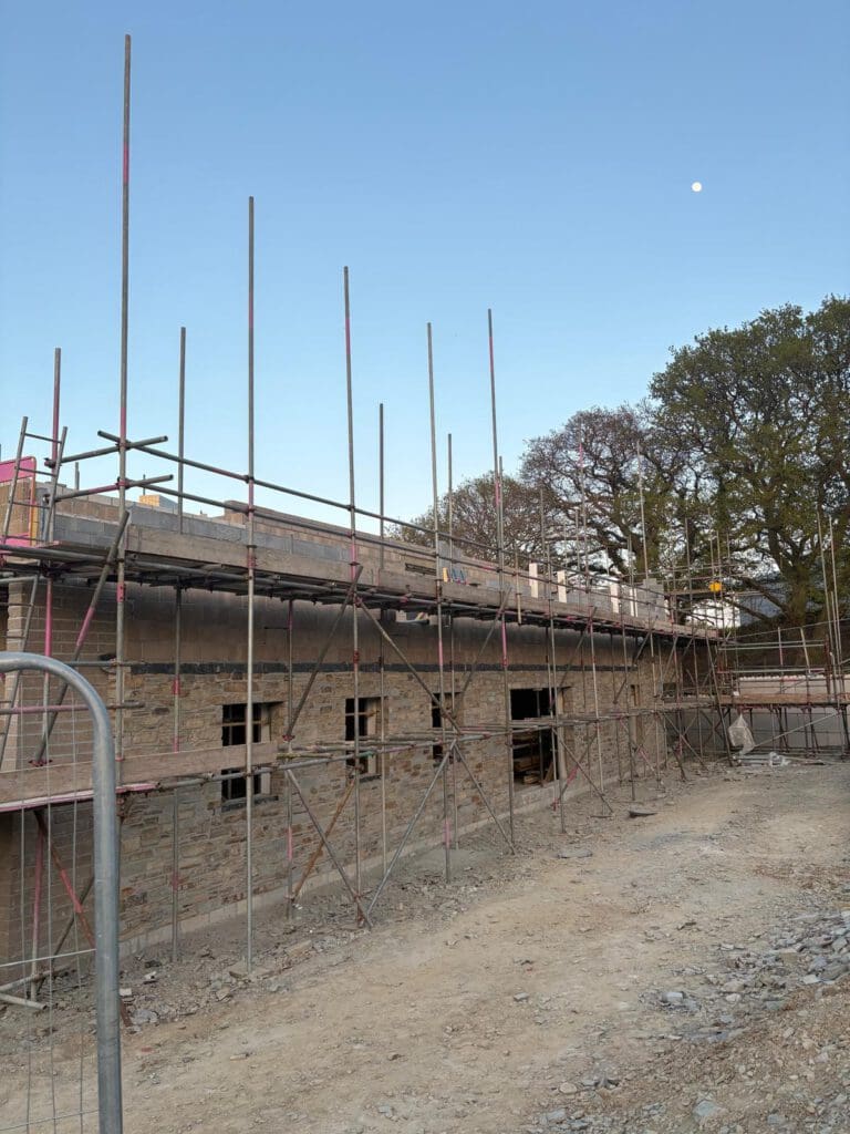 A brick building under construction, surrounded by commercial scaffolding. The ground is bare and uneven, while a clear sky with the moon visible arches above. Trees provide a natural backdrop to the busy work of each skilled scaffolder.