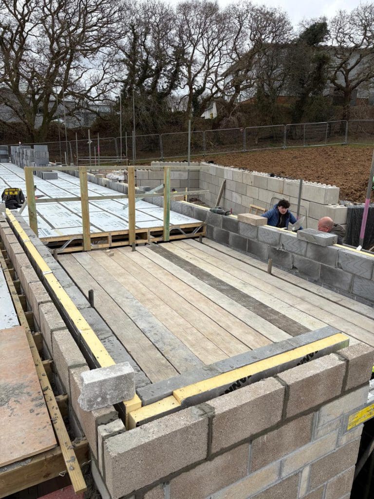 A construction site showing a partially built structure with concrete block walls, wooden planks used as flooring, commercial scaffolding in place, and two workers in blue clothing working on the block walls. Leafless trees and fencing are visible in the background.