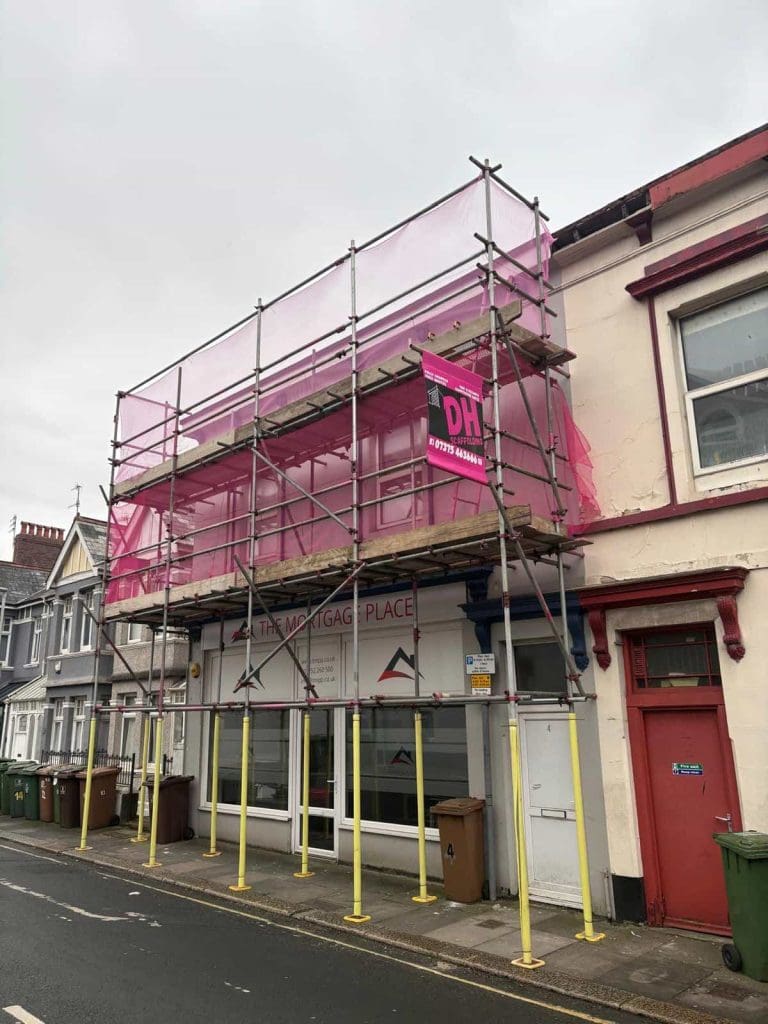 A building with scaffolding and pink safety netting covers the front. A pink sign with white text hangs on the scaffolding. Multiple green bins line the sidewalk beneath the structure. The sky is overcast.